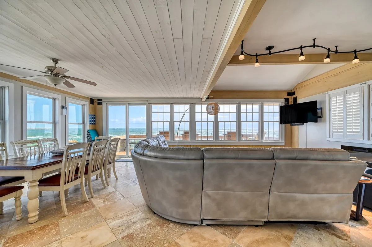 A spacious great room with a plush sectional sofa facing an expansive window showcasing sweeping ocean views. The room includes a large dining table adjacent to the living space, and a wall-mounted television, all flooded with natural light. The open-concept design connects the living and dining areas seamlessly.