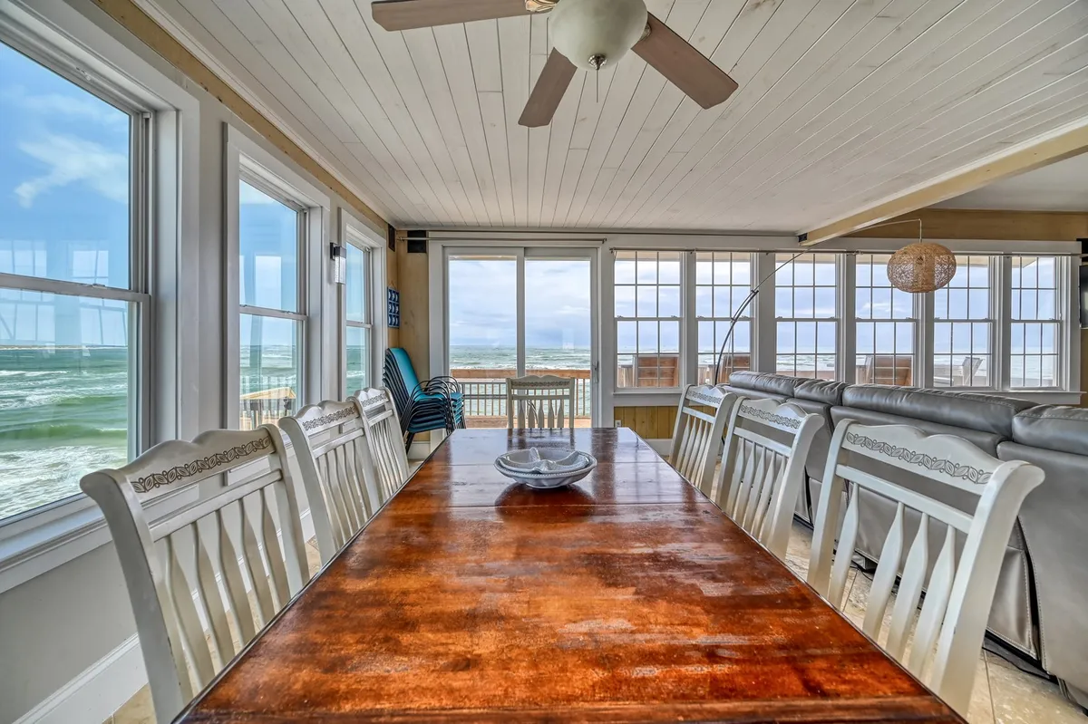 A generous dining area with a large table positioned beside windows that frame the ocean beyond. The room features a ceiling fan and a decorative light fixture, creating an inviting atmosphere for group meals and celebrations while enjoying the coastal view.