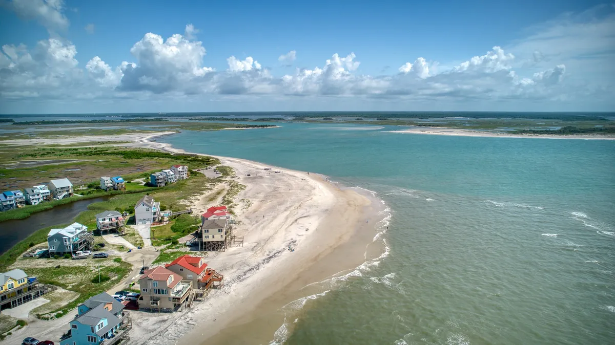 A sweeping view of beachfront homes, a sandbar, and the open ocean under a serene sky. The vibrant yet peaceful community atmosphere and private beach access are highlighted, suggesting opportunities for beachcombing, fishing, water sports, and general relaxation by the sea.