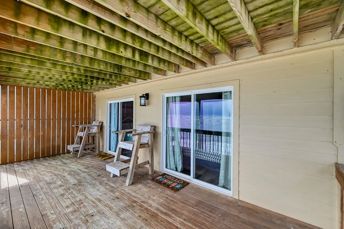 An exterior shot showing a wooden deck with two welcoming chairs, sliding glass doors, and a decorative welcome mat, suggesting a friendly arrival point and connection from the parking area to the home.