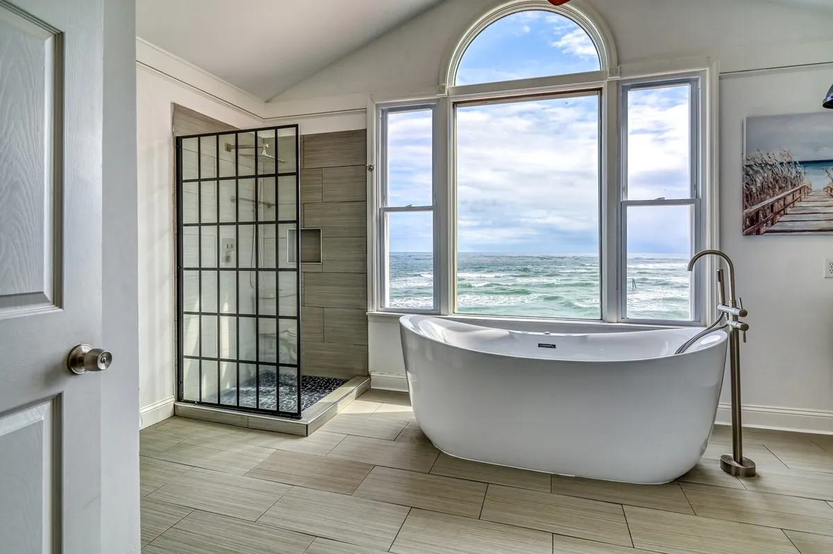 A serene bathroom features a freestanding bathtub positioned next to a large window that frames expansive ocean views, with a sleek glass shower stall nearby. Soft natural light fills the room, emphasizing its calming, spa-like ambiance.