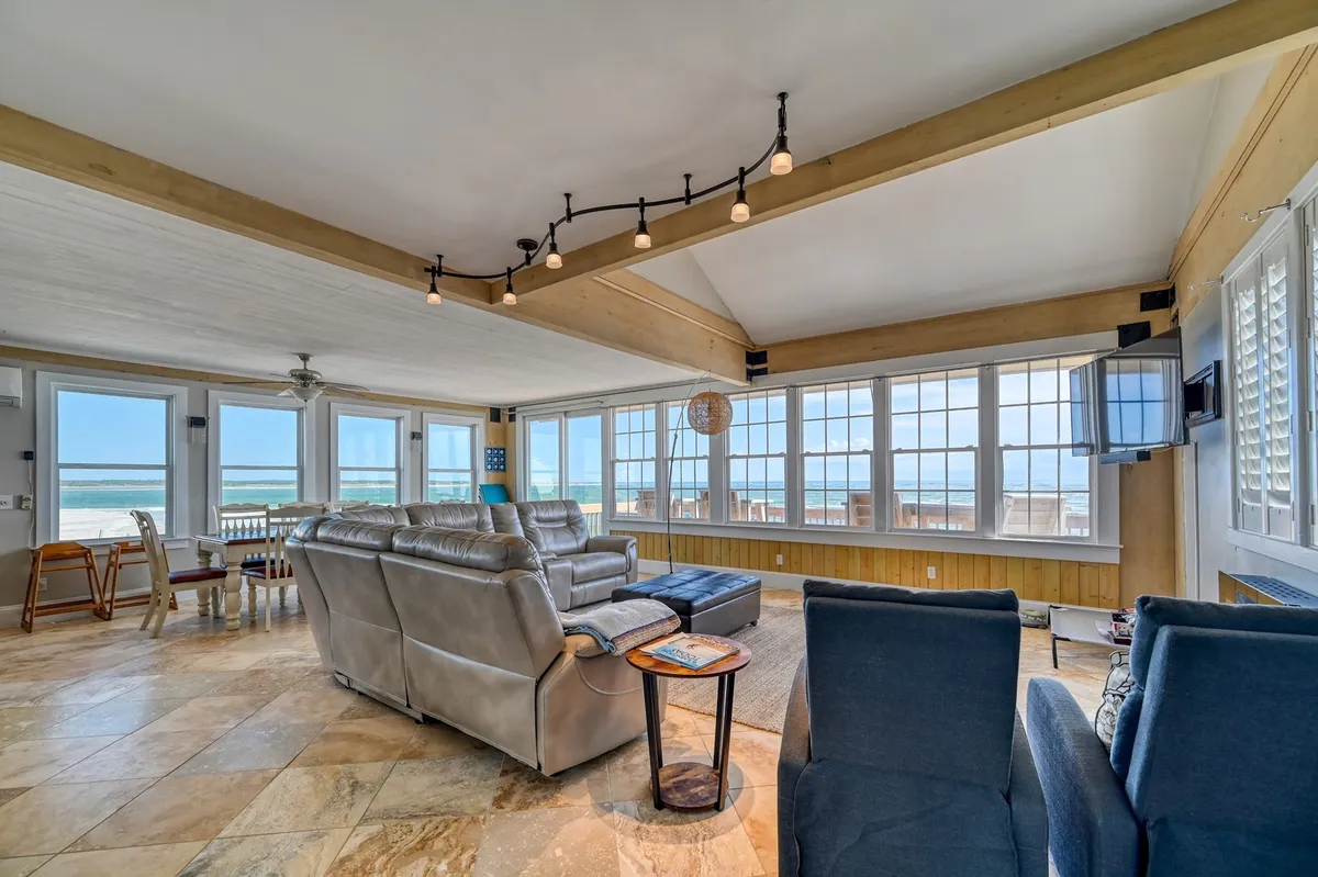 A spacious, bright interior living area featuring large windows framing the ocean, comfortable seating, and inviting decor. The space is set for family movie night or relaxing together indoors after a day at the beach.