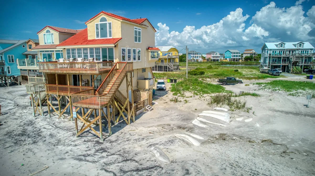 A cheerful exterior view of Kokomo, featuring an elevated beach house with stairs leading up to the entrance. The house is surrounded by colorful neighboring homes, with the sandy beach just steps away, inviting both people and pets for easy access to the shore.