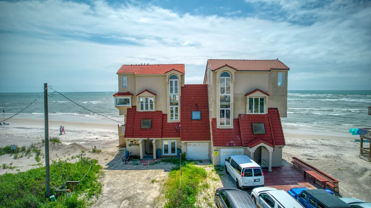 The image showcases a stunning beachfront single-family home with a distinctive red-tiled roof, expansive windows, and two spacious decks overlooking the ocean. The property sits directly on the sand, offering unparalleled access to the beach, and evokes a tranquil, inviting atmosphere ideal for relaxation and gatherings.