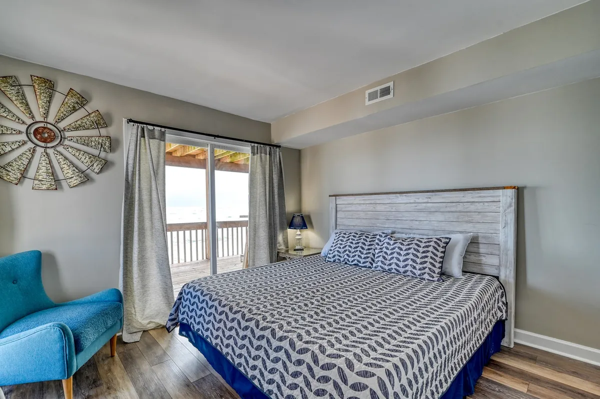 A bright, comfortable bedroom with a patterned bedspread, blue armchair, and a large window dressed with curtains. The room feels peaceful and inviting, with natural light streaming in and a view outside.