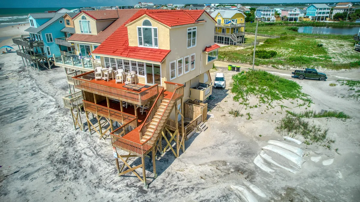 This image captures a vibrant multi-level wooden deck overlooking the beach, with colorful beach houses in the background, sand dunes, and a sweeping ocean view. The deck is a lively space perfect for sunbathing, family gatherings, and direct access to the sand.