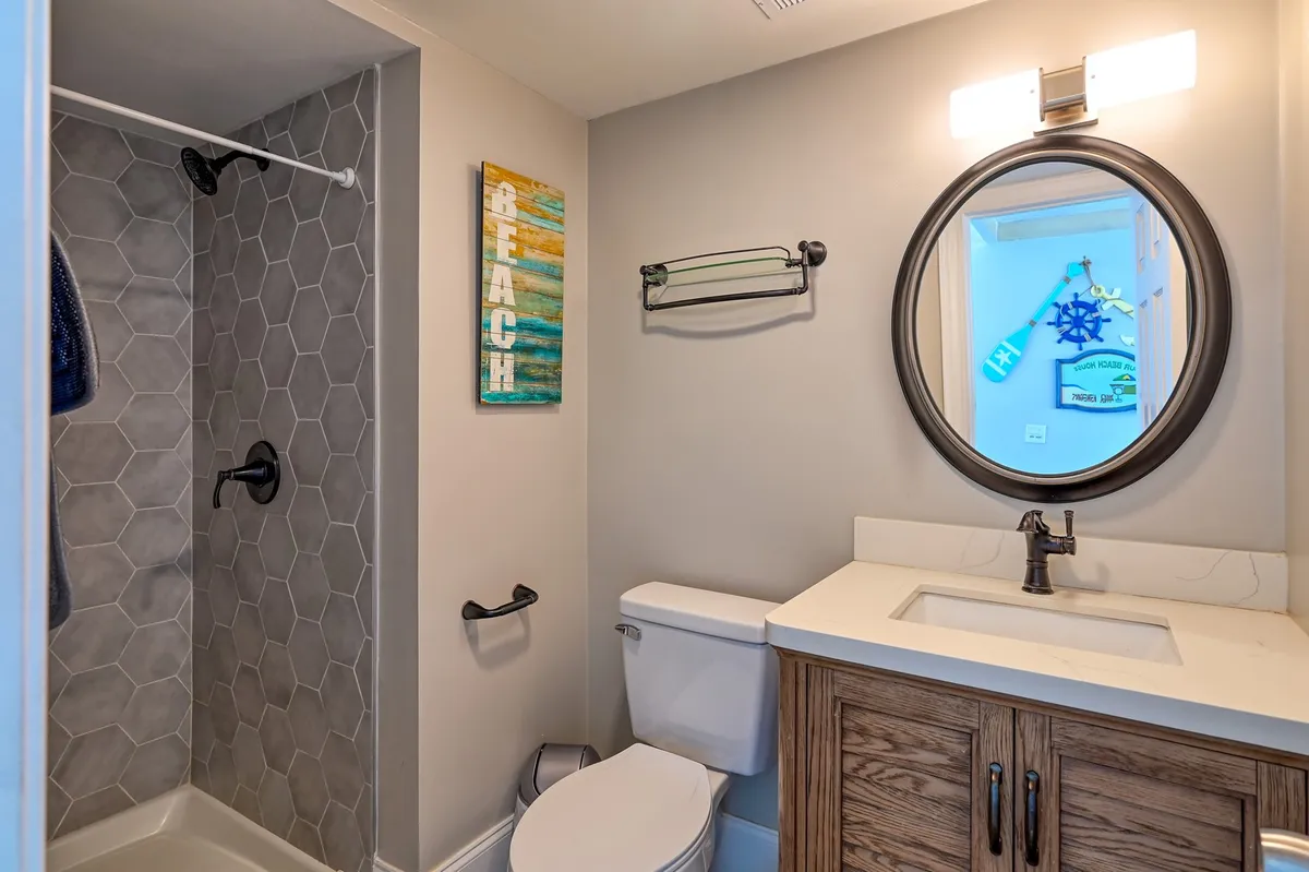 A stylish, hexagonal-tiled shower wall, a round mirror with a black frame, and a wooden vanity define this bright, modern shower space. Beach-themed wall art and refreshing design evoke the ease of rinsing off after a day outside.