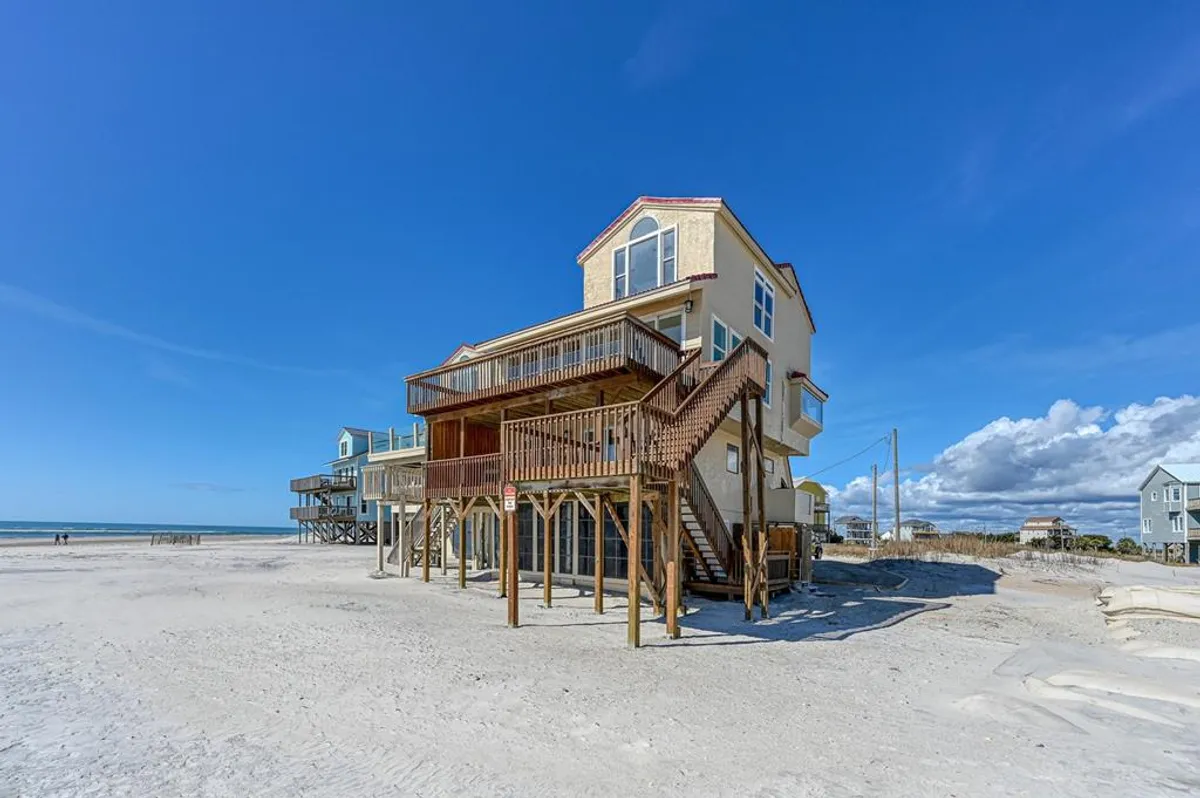 A wide exterior shot of the property with a deck, staircase, and adjacent sandy beach. The scene invites sunbathing, outdoor dining, or walks along the shore, embodying the relaxed lifestyle offered at Kokomo for guests and their pets.