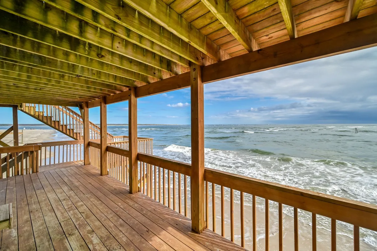 This image showcases a spacious wooden deck overlooking the ocean, with a staircase leading down towards the shore. The scene captures the tranquility of relaxing on the deck, sunbathing, or watching the waves, while also providing access for outdoor activities like sea fishing. The open-air design emphasizes the close connection to the ocean and outdoor recreation.