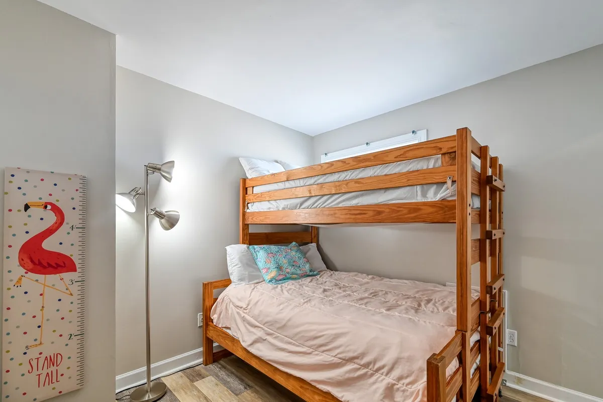 A playful bunk room features a sturdy bunk bed, a flamingo height chart on the wall, a reading lamp, and decorative pillows. The space is styled for children, inviting bedtime stories and quiet play.