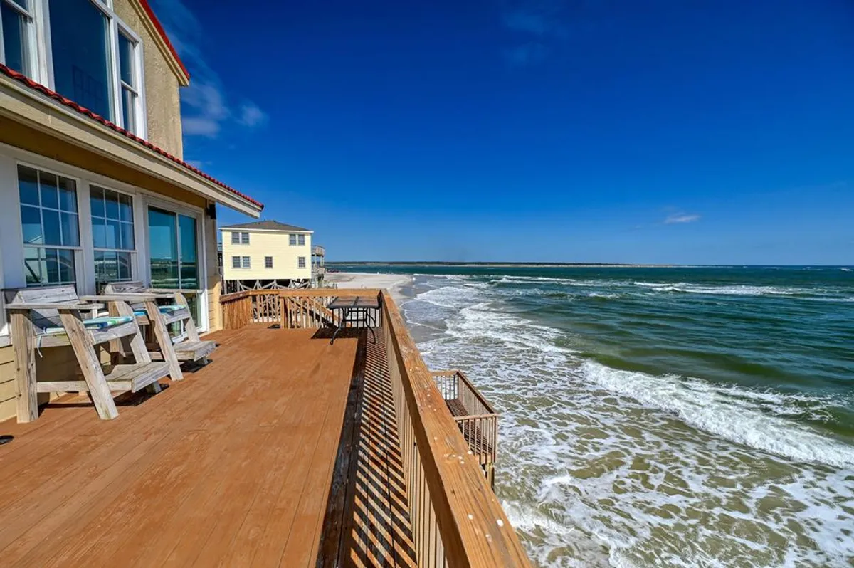 A serene deck directly overlooking the ocean, with waves crashing nearby and neighboring beach houses visible. The open setting is ideal for peaceful moments, sunset dinners, or simply watching the waves roll in.
