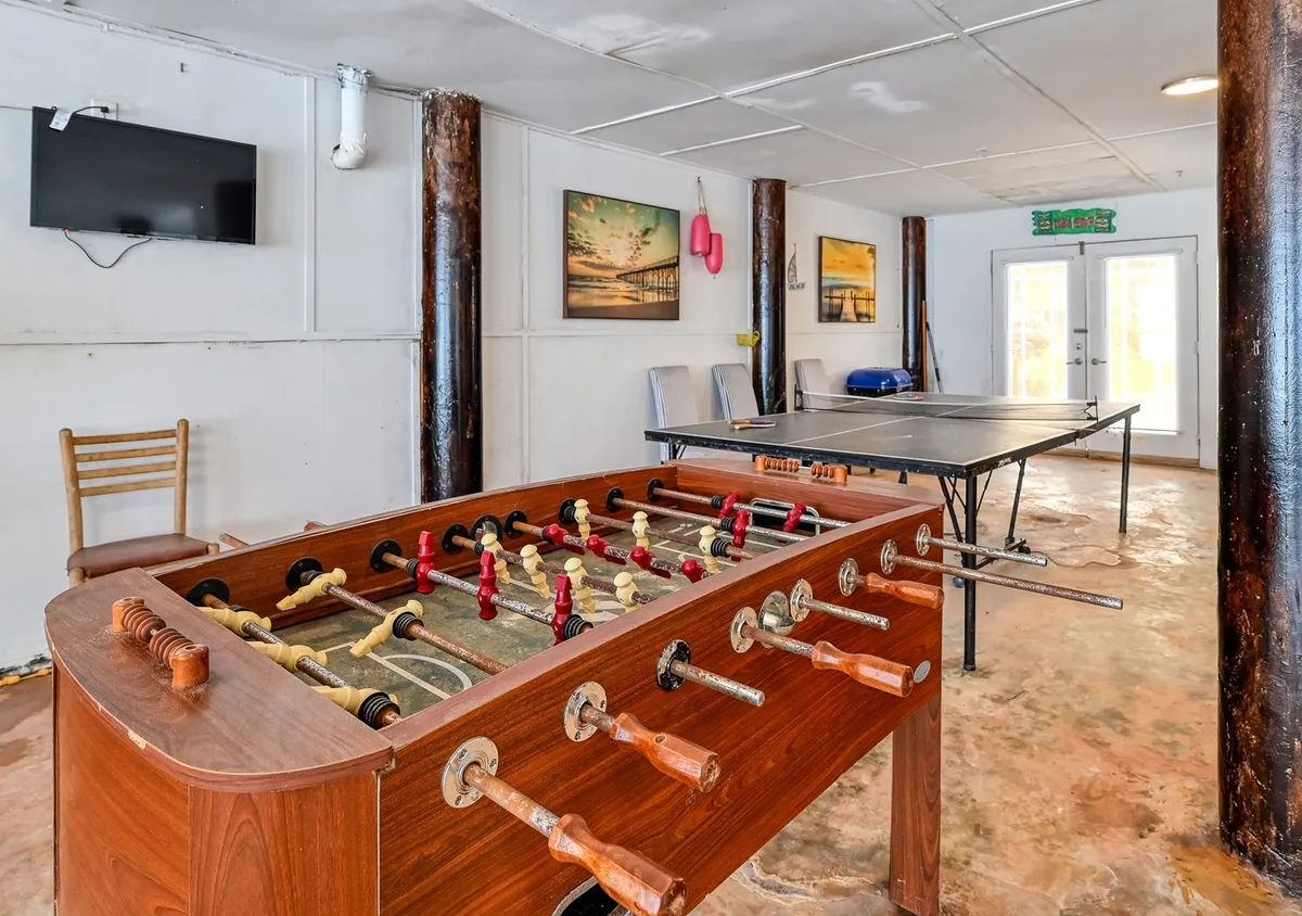 The image shows a lively, well-lit game room featuring both a foosball table and a ping pong table. The walls are adorned with colorful artwork, and a TV screen sits nearby, creating an inviting space for friendly competitions and social gatherings. The room has a relaxed, playful vibe and is clearly designed for group activities and fun.