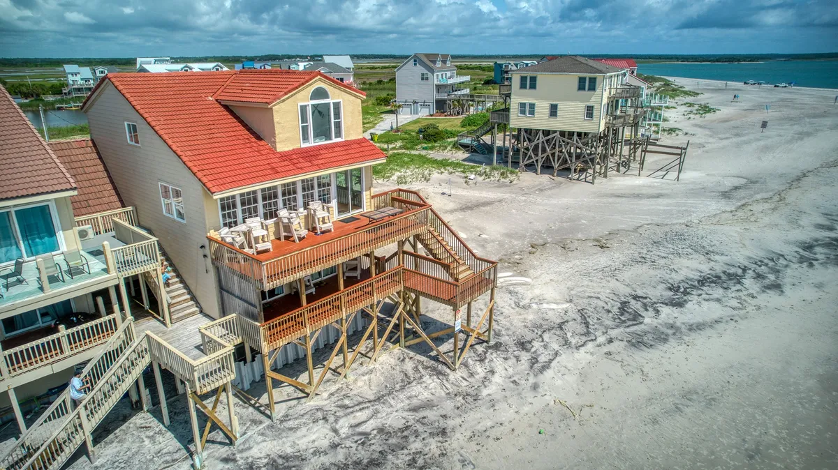 A relaxed exterior image showcasing the beachfront deck of a stilted house overlooking the ocean and sandy beach. The open deck provides plenty of space for people and pets to lounge, play, and soak up the ocean breeze.